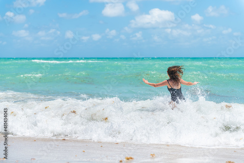 Back of young woman in swimsuit with waves splashing crashing in Sunny Isles, Miami Beach, Florida with Atlantic ocean sea green turquoise water and sargassum seaweed photo