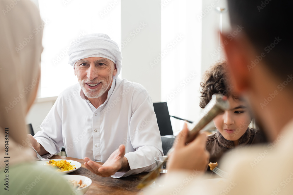 Obraz premium smiling muslim man pointing with hand during dinner with arabian grandson and blurred family.