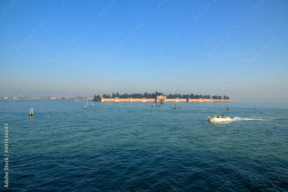 Ein Boot fährt auf dem Türkisblauen Wasser der Lagune von Venedig und im Hintergrund ist bei blauem Himmel die Insel San Michele mit dem gleichnamigen Friedhof zu sehen.