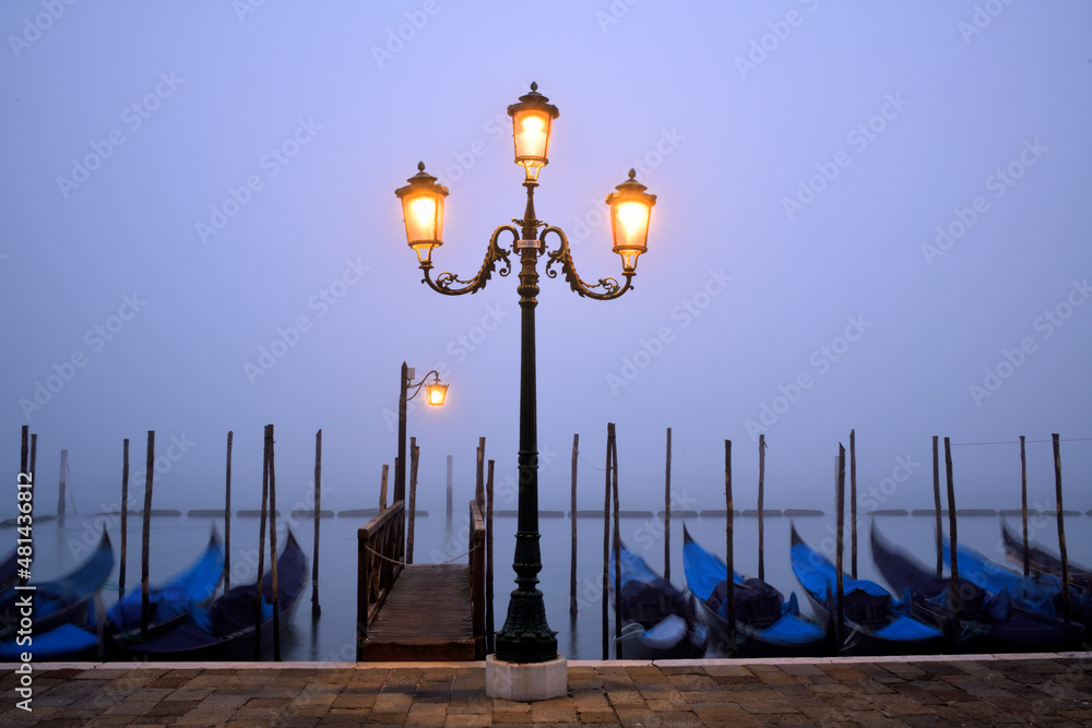 Die Laternen an der Promenade am Canale Grande in Venedig leuchten schon und die Gondeln liegen im Wasser bei einem Nebel verhangenen Hintergrund