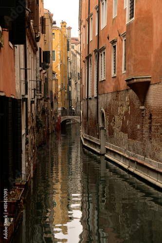 Ein Wasserkanal zwischen typischen Häusern und Palästen von Venedig bei Nebligen Wetter. Weiter weg quert eine Brücke den Kanal und zwei Personen laufen gerade darüber hinweg.
