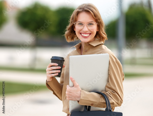 Young cheerful businesswoman walks outside with beaming smile while holding cup of coffee and laptop