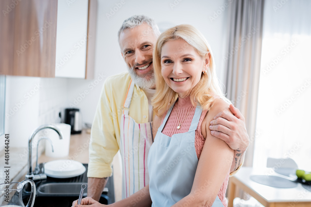 Cheerful mature couple in aprons looking at camera in kitchen. Cheerful mature couple in aprons looking at camera in kitchen.