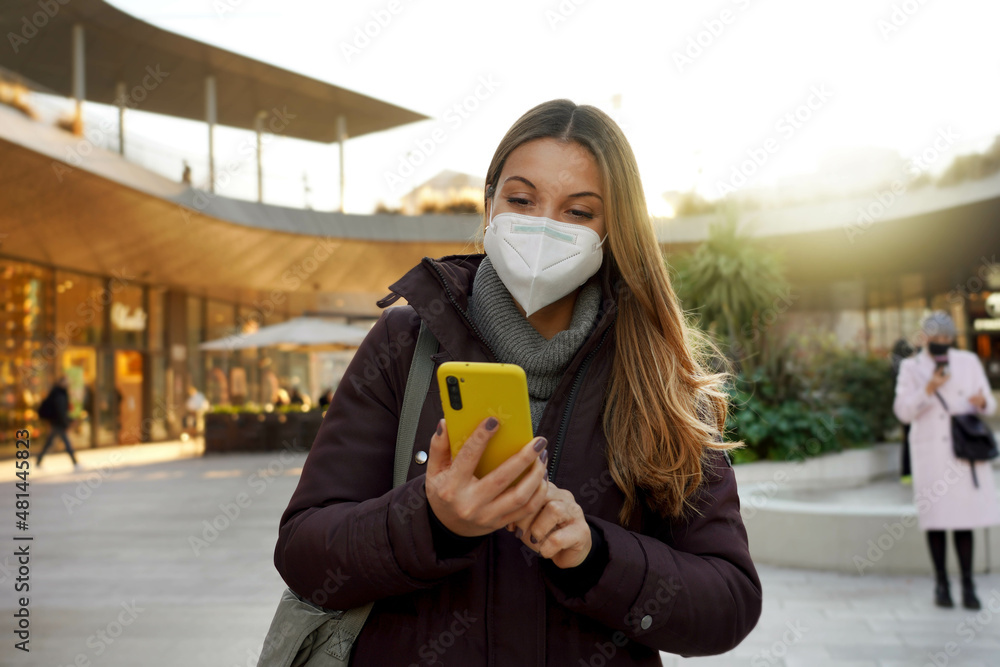 Plakat Portrait of beautiful woman wearing medical face mask using internet wifi on phone in city at sunset