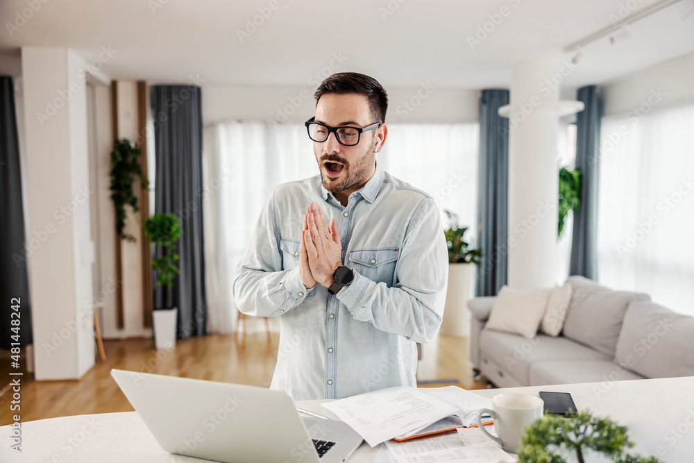 A man standing at his cozy home and praying for good news while looking at the laptop. A man standing at his cozy home and praying for good news while looking at the laptop.