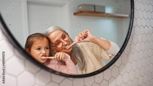 Senior grandmother with granddaughter standing indoors in bathroom, brusing teeth and looking at mirror. photo