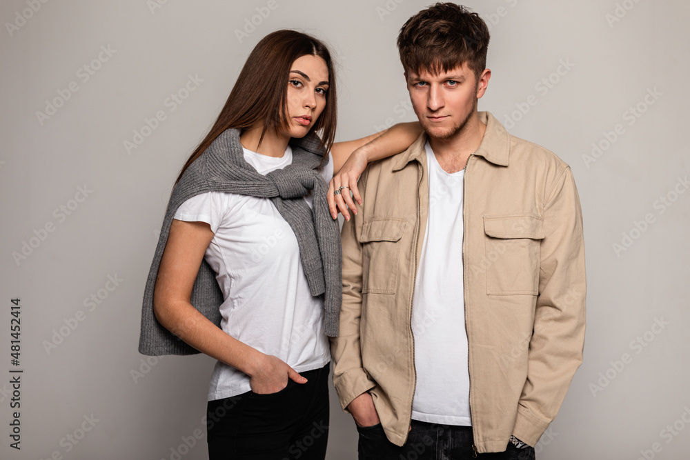Beautiful young stylish couple in trendy casual wear with white t-shirts, knitted sweater and brown jacket stands in the studio. Pretty woman and handsome man in fashionable outwear Beautiful young stylish couple in trendy casual wear with white t-shirts, knitted sweater and brown jacket stands in the studio. Pretty woman and handsome man in fashionable outwear