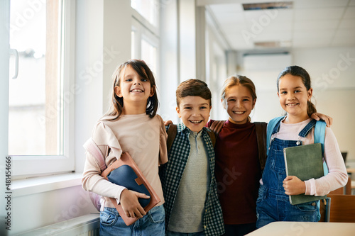 Happy elementary students stand hugged in the classroom and look at camera.