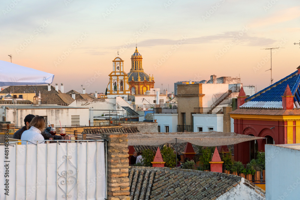 An unidentifiable couple dines outdoors on a rooftop patio in the Barrio Santa Cruz area of Seville, Spain at sunset.