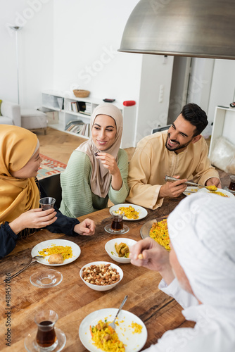 Canvas Print mature asian woman holding glass of tea while talking to muslim family during dinner