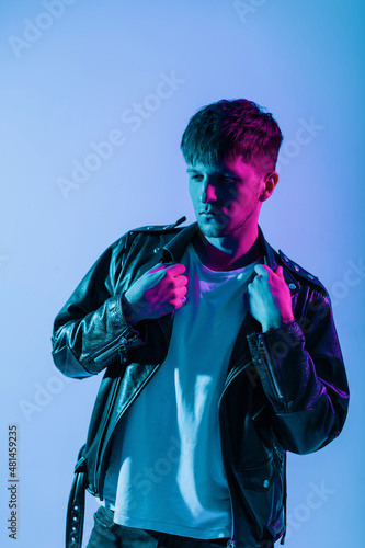 Cool brutal handsome young man with a fashionable hairstyle in a stylish leather retro jacket posing in the studio on neon pink light