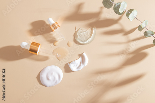 Pipettes and smears and drops of various cosmetic products top view on beige background with dry eucalyptus. Flat lay photo