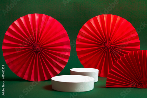 White podium or pedestal with decorative red paper fans on a green background. Product presentation concept. copy space