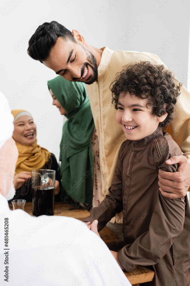 Smiling arabian man hugging son near tea and multiethnic family at home. Smiling arabian man hugging son near tea and multiethnic family at home.