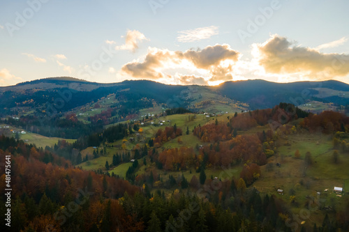 Aerial view of distant village with small shepherd houses on wide hill meadows between autumn forest trees in Ukrainian Carpathian mountains at sunset