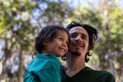 Photography Latin American dad carries his 3 year old son in his arms in the park