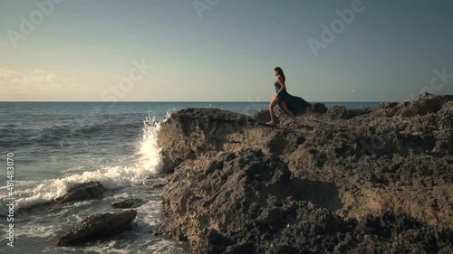 Vista lateral de una hermosa mujer con cabello rizado, con vestido azul caminando sobre las rocas al amanecer en la playa,, disfrutando del viento y la vista
