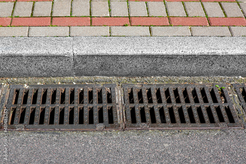 catch basin grate of the lattice of the drain system for drainage of rainwater from the road surface with a tarmac road at the curb with a stone brick tile sidewalk close-up, nobody.