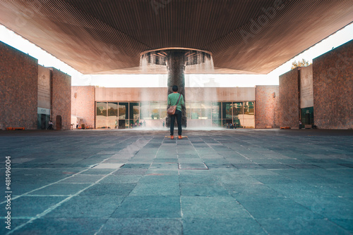 Hombre joven latino, universitario de pie, de espaldas, con una mochila frente a una fuente, dentro de la escuela, vista trasera, de vuelta a la escuela