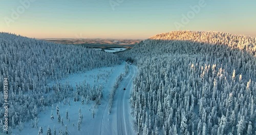 Aerial drone view following a car driving in sunlit, snow covered forest of Lapland photo