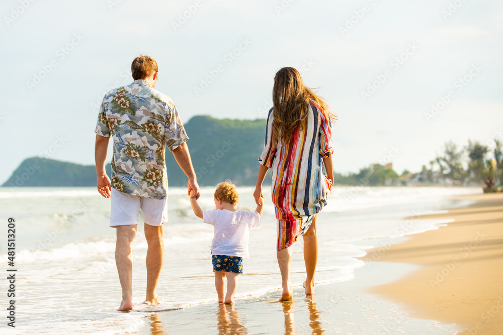 Caucasian couple with cute baby son holding hands walking together on tropical beach. Happy family parents with little child boy enjoy and having fun outdoor lifestyle on summer travel beach vacation.