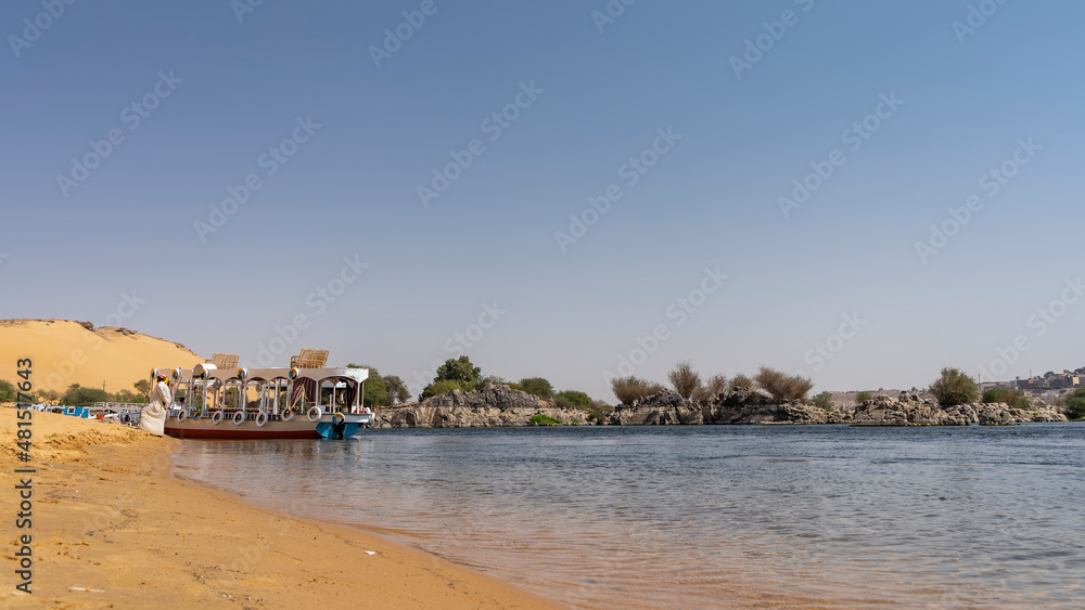 There are tourist boats on the sandy shore of the Nile. Yellow sand of the beach. Ripples on a calm transparent river. In the distance - picturesque boulders, green vegetation. Blue sky. Egypt. Aswan