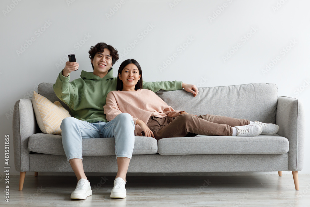 Young Asian couple relaxing on sofa, watching TV, switching channels with remote control, enjoying movie at home