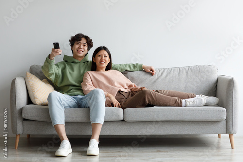 Young Asian couple relaxing on sofa, watching TV, switching channels with remote control, enjoying movie at home