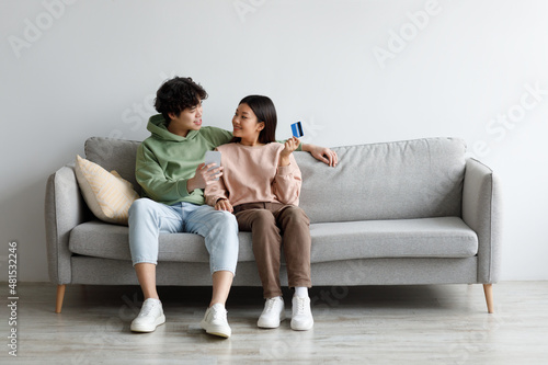 Young married Asian couple with credit card and smartphone sitting on couch, buying in internet store at home