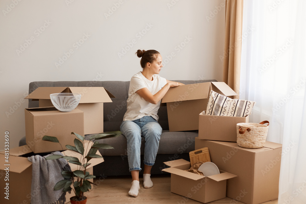 Fototapeta premium Indoor shot of concentrated Caucasian woman wearing white t-shirt and jeans sitting on sofa in a new house or flat after relocating, unpacking her belongings from cardboard boxes.