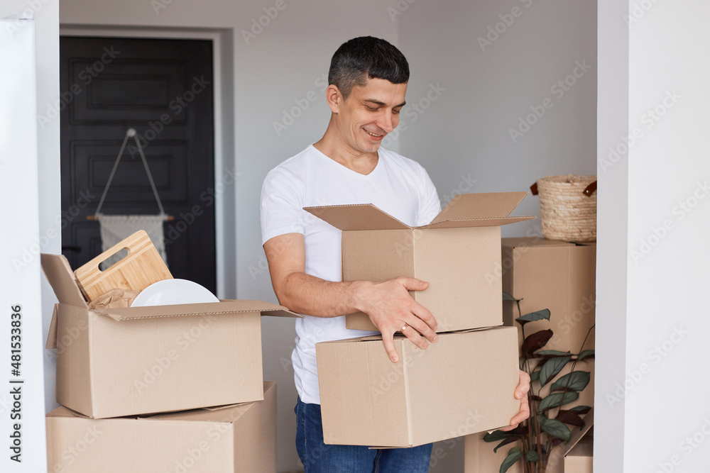 Portrait of optimistic handsome man wearing white T-shirt standing with cardboard boxes with personal stuff, looking inside with happy expression, relocation, real estate. Portrait of optimistic handsome man wearing white T-shirt standing with cardboard boxes with personal stuff, looking inside with happy expression, relocation, real estate.