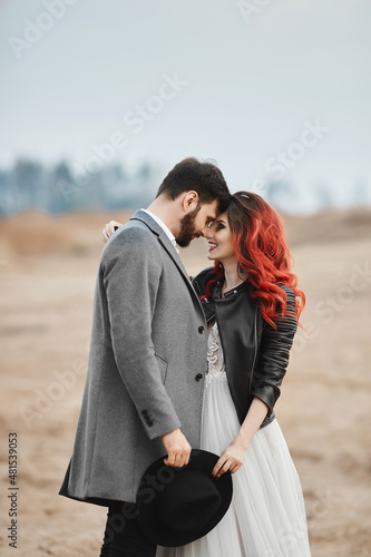 A fashionable couple of newlyweds posing outdoors. Stylish bearded man and his red-haired girlfriend in a wedding dress and leather jacket © innarevyako