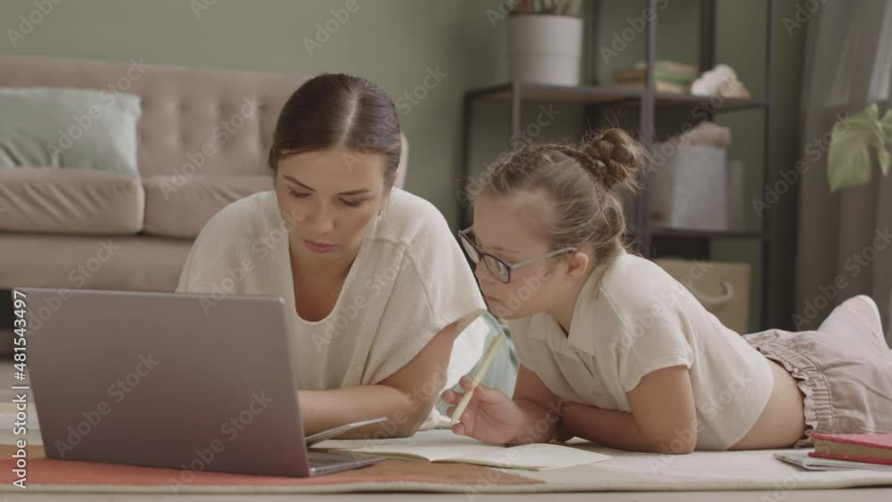 Slowmo shot of mom and daughter studying on laptop together lying casually on floor in living room Slowmo shot of mom and daughter studying on laptop together lying casually on floor in living room