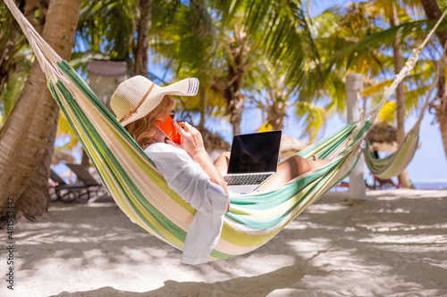 Woman lying in hammock on tropical beach and working on laptop and speaking on phone photo