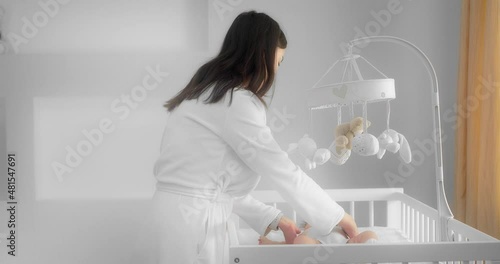 Mother in a white coat picks up a crying newborn baby lying in a crib and calms him down. Infant in the white nursery. Concept of Childhood, New Life, Parenthood. Shot with RED camera