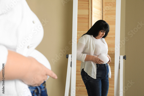 Overweight woman in tight shirt near mirror at home photo
