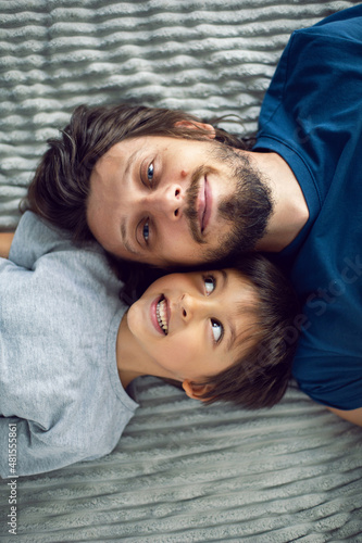 bearded father and his son are lying on the bed with phone and taking selfies at the window in a white room