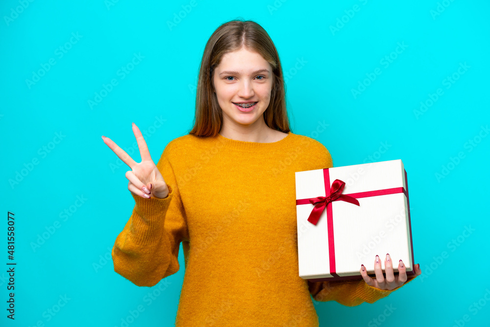 Teenager Russian girl holding a gift isolated on blue background smiling and showing victory sign Teenager Russian girl holding a gift isolated on blue background smiling and showing victory sign