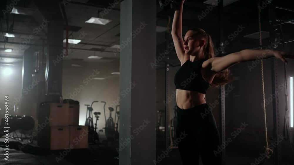 A strong woman makes efforts and overcoming difficulties lifts a dumbbell in a dark gym. Fitness woman lifting weight dumbbells training in gym club. A strong woman makes efforts and overcoming difficulties lifts a dumbbell in a dark gym. Fitness woman lifting weight dumbbells training in gym club.