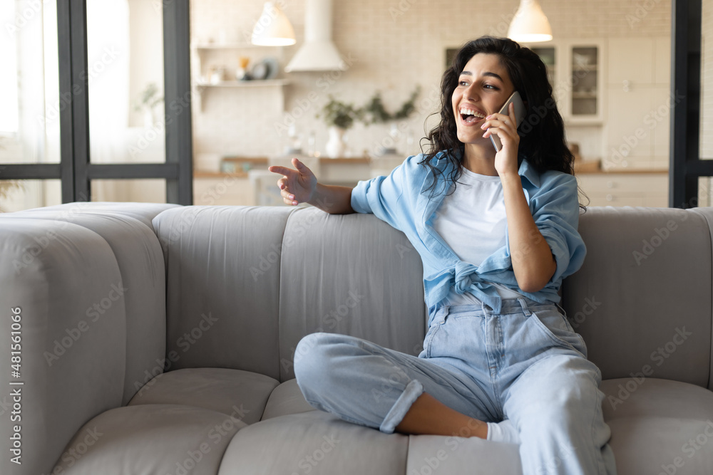 Cheerful young brunette lady sitting on sofa, making call on mobile phone at home, copy space