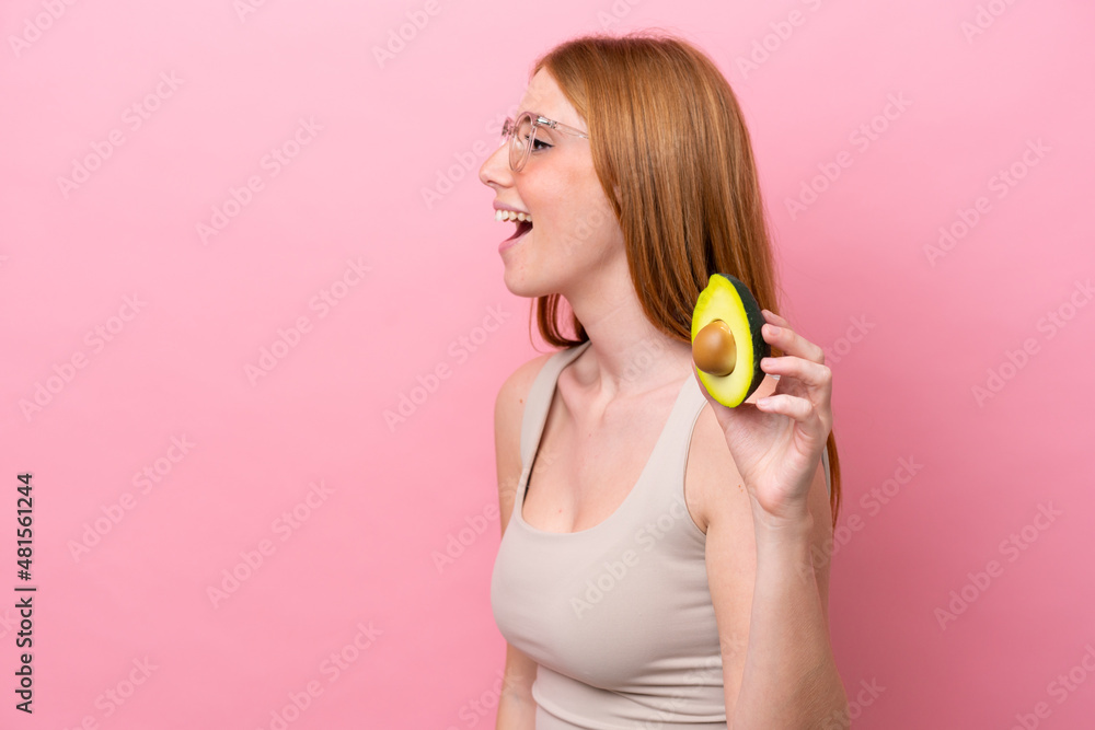 Young redhead woman holding an avocado isolated on pink background laughing in lateral position Young redhead woman holding an avocado isolated on pink background laughing in lateral position