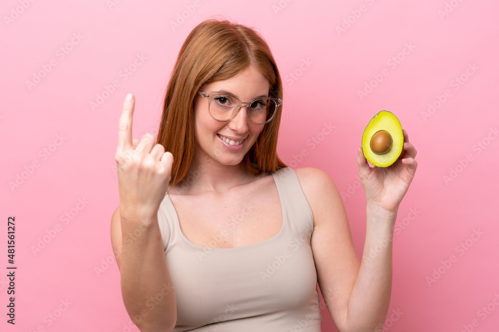 Young redhead woman holding an avocado isolated on pink background doing coming gesture Young redhead woman holding an avocado isolated on pink background doing coming gesture