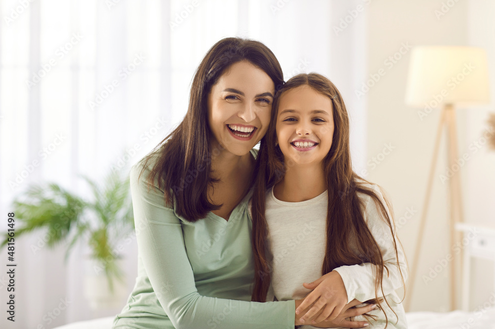 Indoor portrait of happy Caucasian mom and child at home. Cheerful beautiful young mother and pretty little daughter sitting together on bed, looking at camera and smiling. Family, love, care concept Indoor portrait of happy Caucasian mom and child at home. Cheerful beautiful young mother and pretty little daughter sitting together on bed, looking at camera and smiling. Family, love, care concept