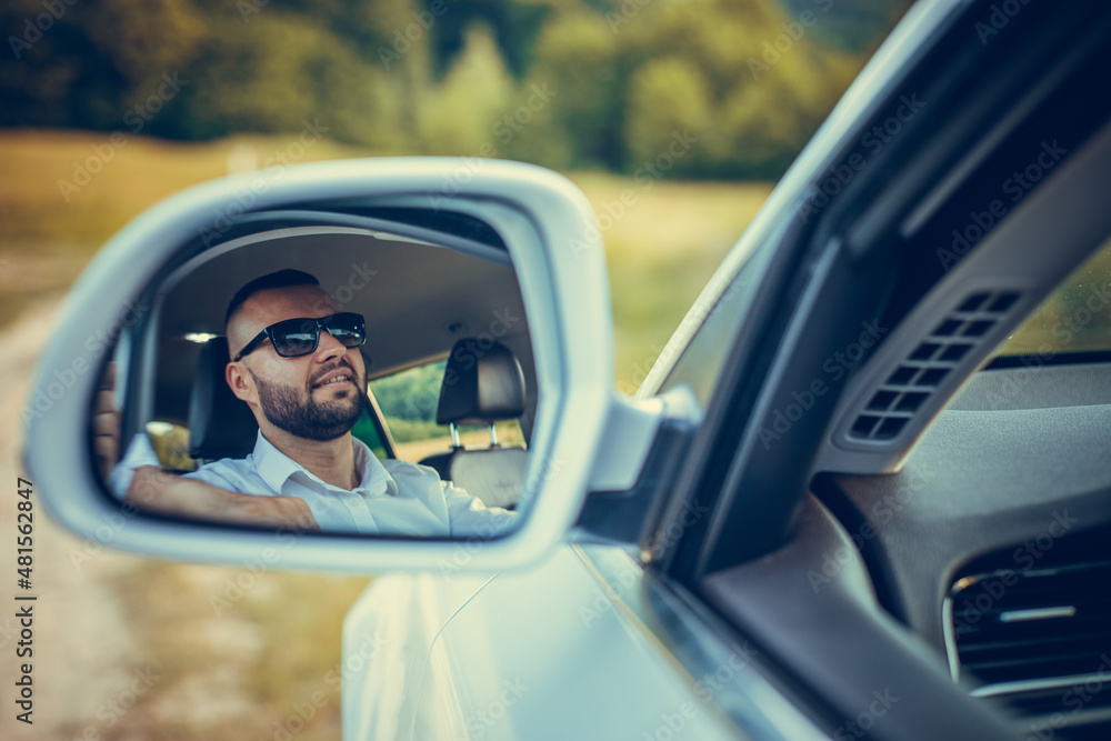 Naklejka premium Bearded caucasian business man looking at camera through rear view mirror. Head and shoulders. Summer background.