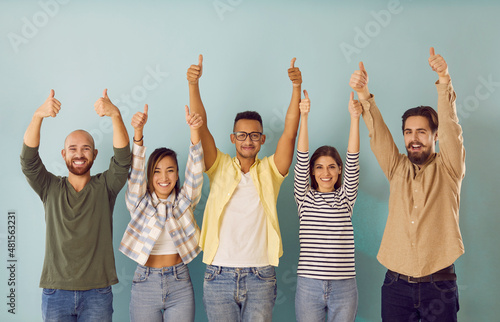 Different ethnic young people make happy gesture by raising their hands with thumbs up demonstrating success. People in casual clothes stand on light blue background. Approval and feedback concept.
