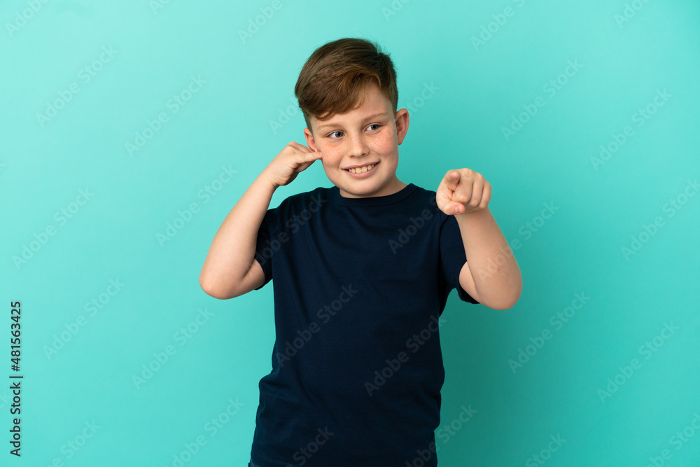 Little redhead boy isolated on blue background making phone gesture and pointing front Little redhead boy isolated on blue background making phone gesture and pointing front