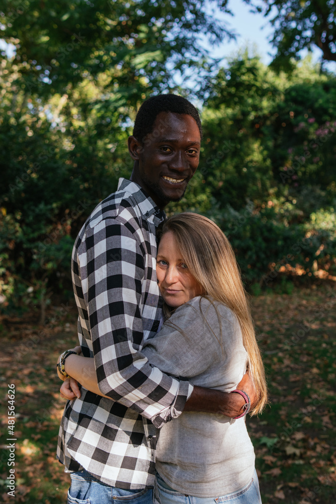 A couple of an African man and white woman are embracing and smiling in public green area A couple of an African man and white woman are embracing and smiling in public green area