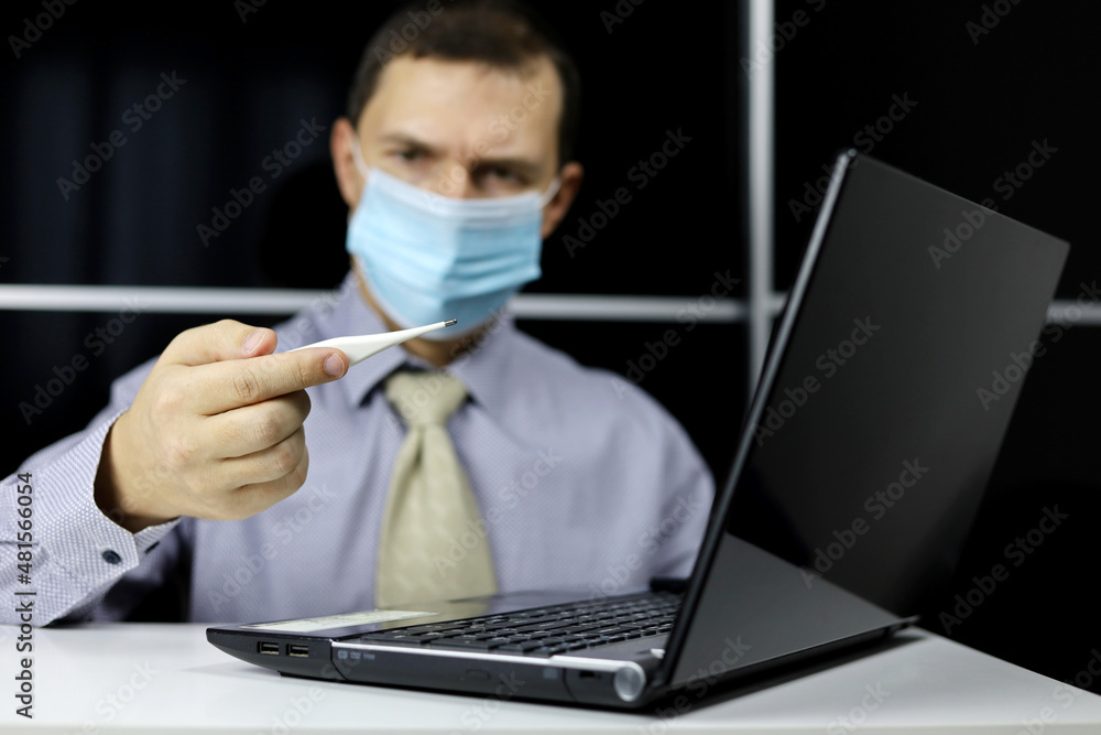 Worried man in face mask and office clothes looks at digital thermometer in hand sitting at laptop. High body temperature, safety at work during coronavirus pandemic Worried man in face mask and office clothes looks at digital thermometer in hand sitting at laptop. High body temperature, safety at work during coronavirus pandemic