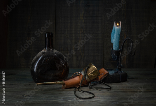 An old dirty bottle of alcoholic beverages, an inlaid horn in a case and a candle holder with a burning candle on a wooden table on a dark background