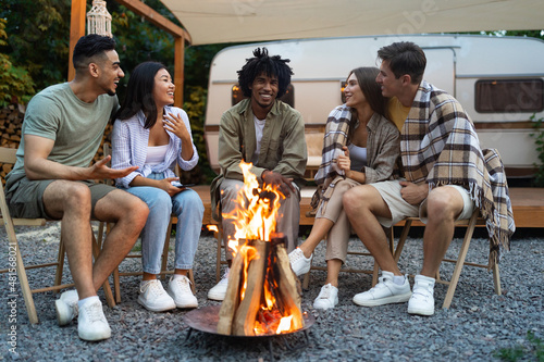 Multiracial friends sitting near bonfire, talking to each other, laughing, spending autumn evening near motorhome